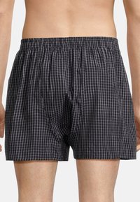 Schwarze Boxershorts mit weißem Karomuster, elastischem Bund und Seitentaschen. Das Material wirkt leicht und atmungsaktiv.