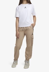 Bílá tričko s krátkým rukávem s černým logem Jordan, kombinované s béžovými cargo kalhotami se bočními kapsami a černými akcenty, obuté s lehkými teniskami.