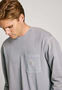 Light grey long-sleeve shirt made of soft fabric, featuring a chest pocket with a label. Round neckline and relaxed fit.