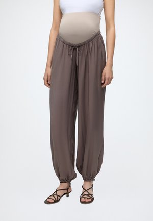 Femme enceinte portant un pantalon de maternité marron large avec des chevilles élastiques et une taille à cordon, associé à des sandales noires à lanières.
