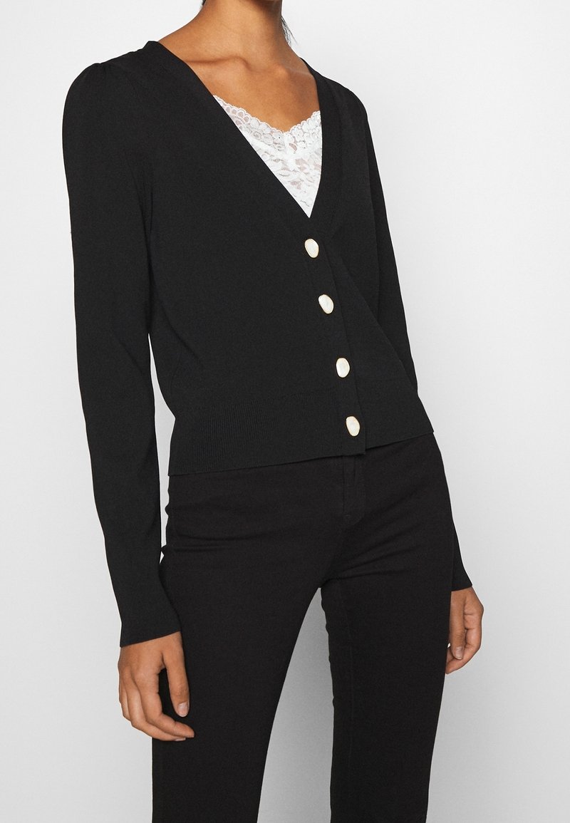 Cardigan noir à encolure en V profonde, manches longues et cinq boutons crème à l'avant. Présente une texture côtelée à l'ourlet et aux poignets.