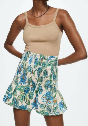 Beige geribbelde tanktop en een korte rok met een bloemenpatroon in blauw en groen op een lichte achtergrond; smocktail en gerimpelde zoom.