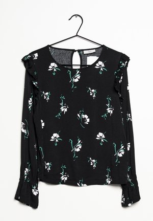 Sort langærmet bluse med hvidt og grønt blomsterprint, flæsekant på ærmerne og skulderdetaljer, hængende på en sort bøjle.