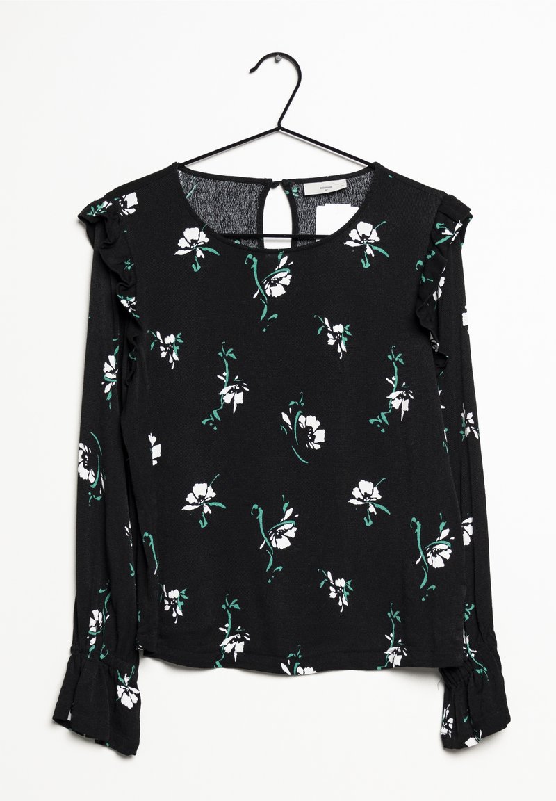Blouse noire à manches longues avec motif floral blanc et vert, poignets à volants et détails aux épaules, suspendue à un cintre noir.