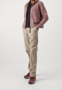 Chaqueta de forro polar de color rosa claro con cuello alto, combinada con pantalones cargo beige que tienen dos bolsillos frontales, y zapatillas deportivas rojas y grises.