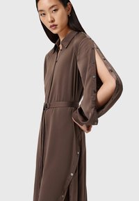 Vestito marrone a maniche lunghe in un tessuto liscio con vita con cintura, caratterizzato da una chiusura frontale con bottoni e spaccature aperte sulle maniche.
