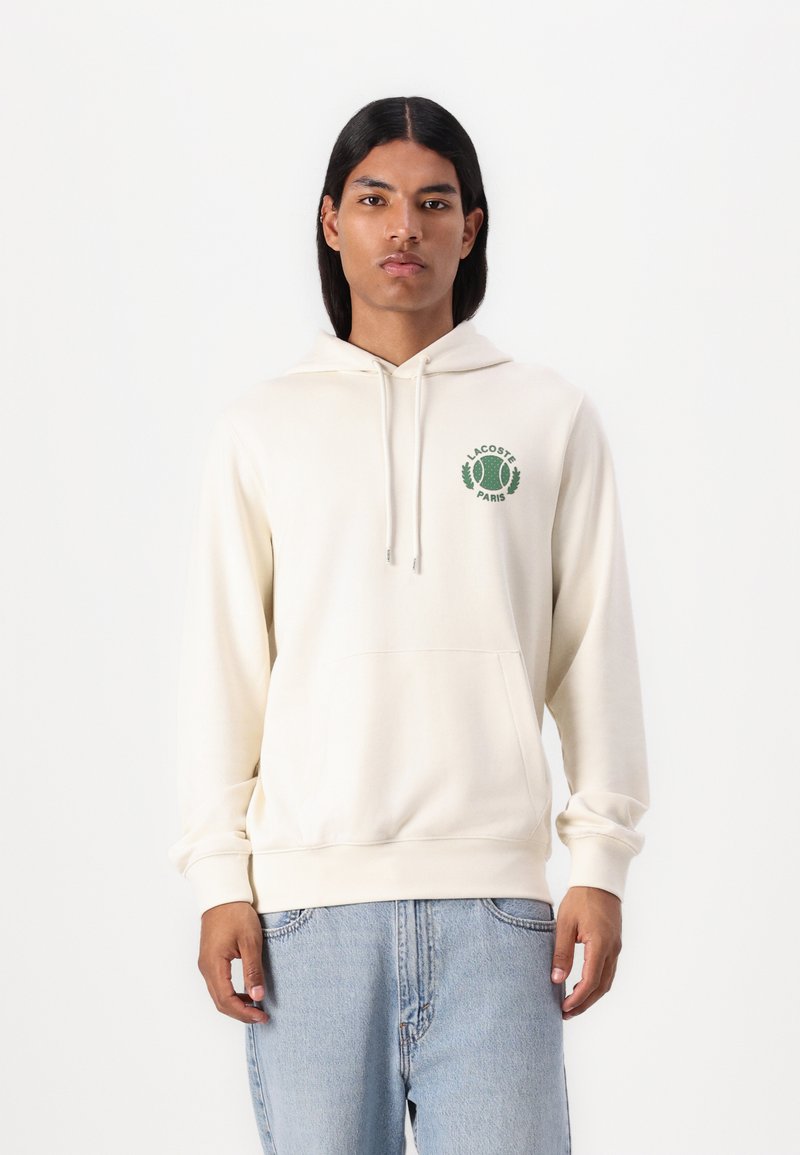 Sudadera con capucha de color beige claro con un bolsillo tipo canguro, bordado verde con motivos de tenis y el texto "LACOSTE PARIS", textura suave.