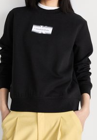 Svart bomullströja med ribbade ärmslut och fåll, med en vit etikett med "Calvin Klein Jeans" i fet stil på bröstet.