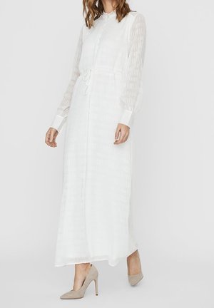 Frau in einem langen, weißen, strukturierten Kleid mit durchsichtigen Ärmeln, geknöpftem Vorderteil, Taillengürtel und beigen High Heels, die vor einem schlichten Hintergrund steht.