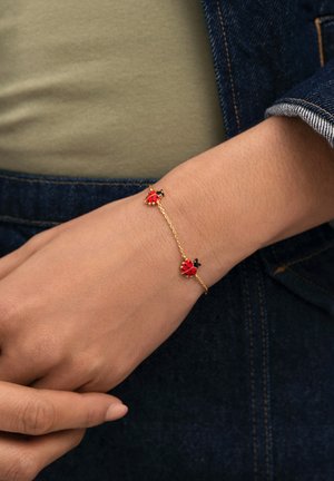 Pulsera de cadena de oro con dos dijes de mariquitas rojas y negras, llevada en la muñeca sobre un fondo de mezclilla y tela verde.