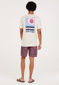 Light beige t-shirt with a colorful printed design on the back featuring waves, sun, and clouds. Paired with patterned pink shorts and sandals.