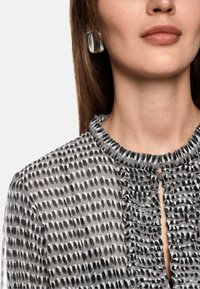 Graue Bluse mit einem Rüschenausschnitt und einer Knopfverschlüsse. Sie hat ein Muster aus schwarzen und weißen Punkten in unterschiedlichen Größen.