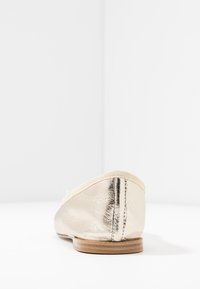 Bailarina metálica dorada con un suave ribete de tela, punta redondeada, textura lisa y suela de madera plana. La vista trasera muestra detalles de costura.