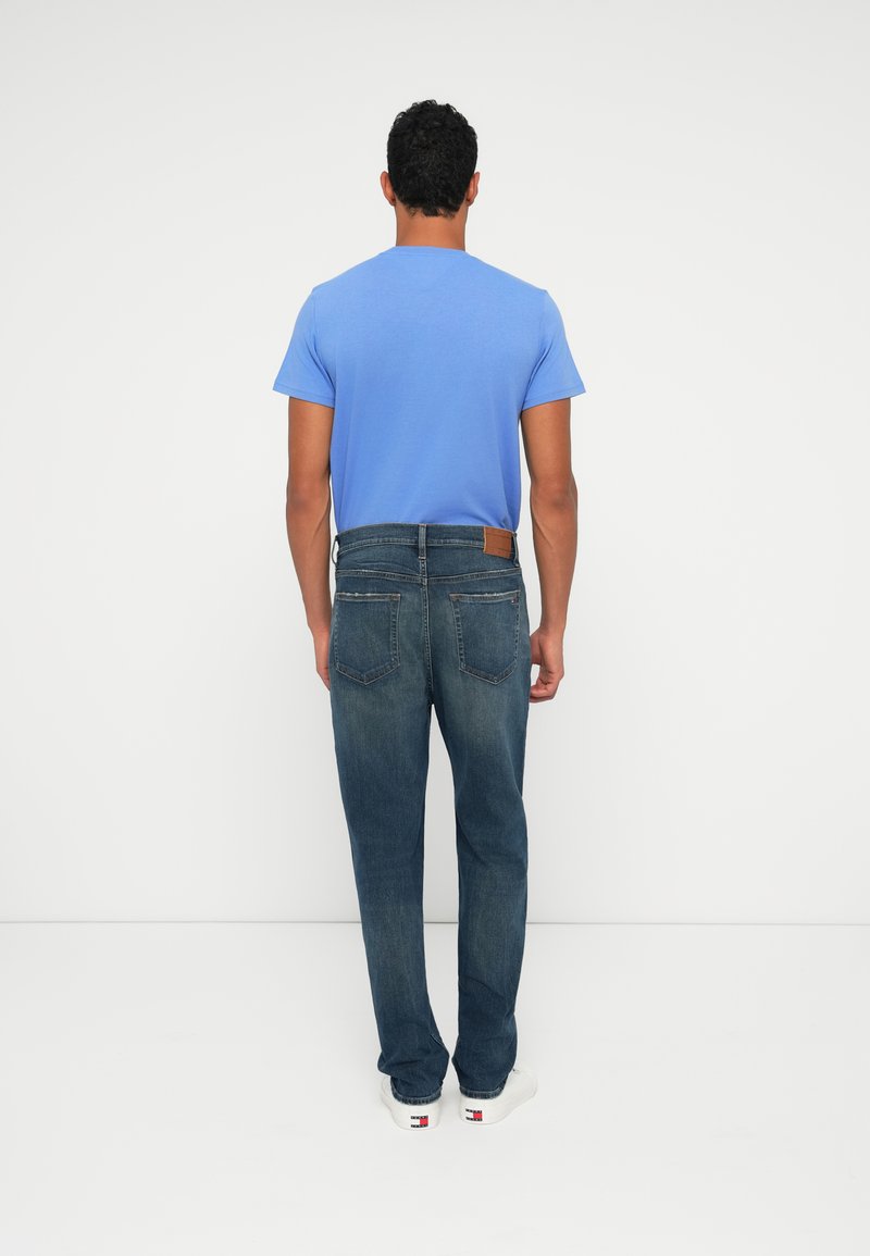 Blue cotton t-shirt paired with dark blue denim jeans. The jeans feature a leather patch and standard back pockets. White sneakers complete the look.