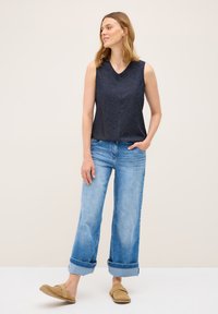 Granatowy top bez rękawów o teksturowanym wzorze, w połączeniu z jasnoniebieskimi spodniami o szerokich nogawkach i beżowymi butami wsuwanymi, stojący na neutralnym tle.