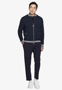 Marineblå strikket bomberjakke med ribbet krave og manchetter, udstyret med en sølvfarvet lynlås og to sidelommer, sammen med marineblå tapered bukser og hvide sneakers.