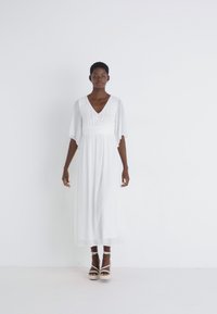 Μακριά λευκή φόρεμα με V-λαιμόκοψη, κοντά μανίκια και εφαρμοστό μέση. Φτιαγμένο από ελαφρύ ύφασμα, ρέει μέχρι το μήκος του μέσου μόσχαλου.