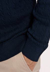 Marinel blauw kabelgebreide trui met geribbelde manchetten en zoom, gecombineerd met beige broeken. De textuur is zacht, met opvallende gedraaide patronen.