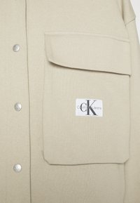 Tissu beige avec grande patte de poche et boutons-pression métalliques embossés avec "CK" sur le côté gauche, avec une étiquette Calvin Klein Jeans.