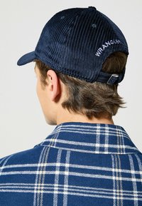 Marinfärgad kordmössa med en böjd skärm, med vit "WRANGLER" broderi på baksidan. Jackan har ett blått och vitt rutmönster.