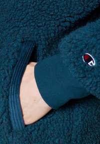 Teal fleece tyg med en mjuk, texturerad yta. Har en ribbad manschett och en ficköppning, samt en logotypdetalj på ärmen.