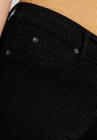 Vaqueros de mezclilla negra con una textura suave, diseño clásico de cinco bolsillos y acentos de remaches metálicos en el bolsillo trasero y la cinturilla.