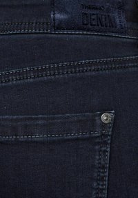 Jeans de mezclilla oscura con una superficie texturizada, que cuentan con un bolsillo trasero, detalles de remaches metálicos y una etiqueta negra con letras en relieve.