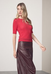 Crveni teksturirani top s kratkim rukavima, uparен s smeđom kožnom suknjom s vertikalnim šavovima. Neutralna pozadina.