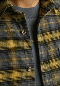 Camicia a quadri caratterizzata da colori grigi, neri e gialli, con una consistenza morbida e colletto rotondo, abbinata a una maglietta grigia a girocollo.