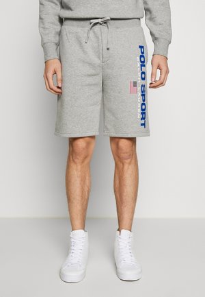 Mann trägt hellgraue Shorts mit "Polo Sport Ralph Lauren" und einem amerikanischen Flaggen-Schriftzug am rechten Bein, kombiniert mit weißen High-Top-Sneakers.