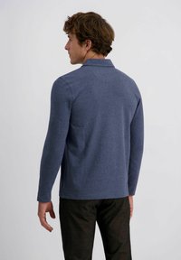 Mann in einem langärmeligen blauen Poloshirt und dunklen Hosen, der leicht zur Seite gedreht vor einem schlichten weißen Hintergrund steht.