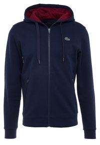 Laivastonsininen vetoketjullinen huppari viininpunaisella vuorella, jossa pieni vihreä logo vasemmassa rinnassa ja etutaskut. Valmistettu pehmeästä kankaasta.