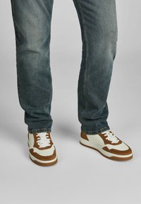 Braune und weiße Sneaker aus Wildleder und Leder mit einer durchbrochenen Zehenpartie, weißen Schnürsenkeln und einer Gummisohle. Getragen mit blauen Jeans.