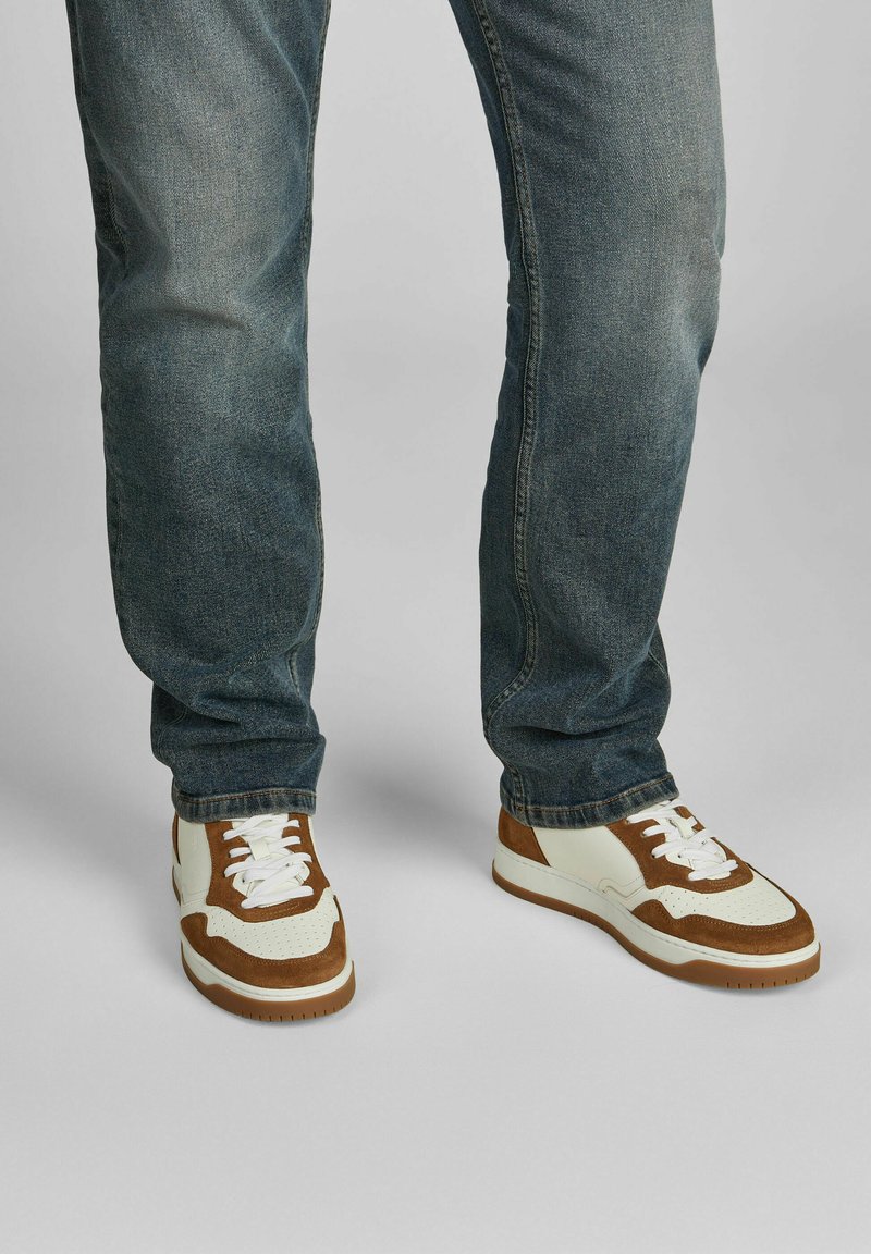 Braune und weiße Sneaker aus Wildleder und Leder mit einer durchbrochenen Zehenpartie, weißen Schnürsenkeln und einer Gummisohle. Getragen mit blauen Jeans.