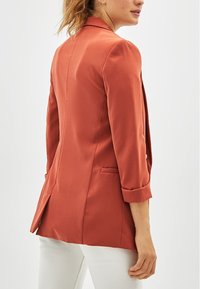 Terakotový blazer s jednopaznovým zapínáním, přiléhavý střih a ohrnuté rukávy. Obsahuje dvě boční kapsy a hladký povrch.