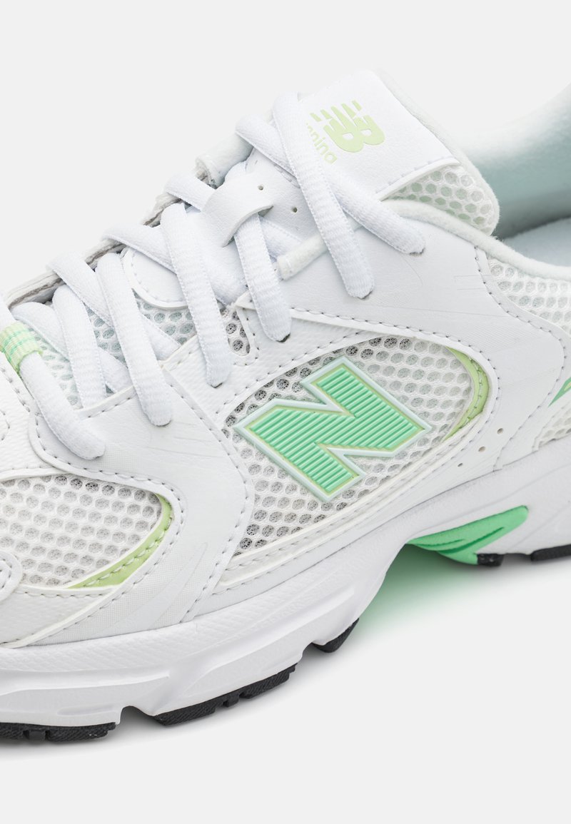 Zapatilla deportiva blanca con paneles de malla, detalles en verde y una suela de goma texturizada. Logo destacado en el costado. Los cordones están asegurados.