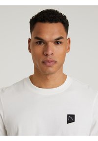 Weißes Baumwoll-T-Shirt mit rundem Halsausschnitt und einem kleinen schwarzen Logo-Patch auf der linken Brust. Glatte Textur, reguläre Passform.