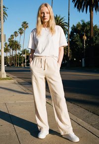 Young woman stands on sunny sidewalk between palm trees wearing a white t-shirt, beige drawstring pants, and white sneakers.