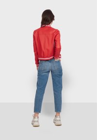 Kobieta stojąca tyłem do aparatu, ubrana w czerwoną kurtkę, niebieskie dżinsy i białe sneakersy, na jednolitym jasnym tle.