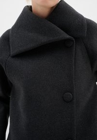 Schwarzer Wollmantel mit großem Kragen, runden Knöpfen und strukturiertem Design, mit glatter Textur und minimaler Ausstattung.