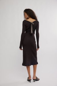 Vestido de malha castanho com mangas compridas, decote em V profundo nas costas e detalhe com laço de fita. Silhueta ajustada e bainha evasê. Usado com sandálias de salto pretas.