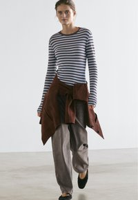 Striped long-sleeve top in blue and grey, brown suede wrap, loose grey pants, and black slip-on shoes. Smooth textures and casual fit.