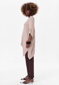 Beige gebreide poncho met een losse pasvorm, voorzien van omgeslagen mouwen, asymmetrische onderkant, gecombineerd met donkerbruine broek en gepatroniseerde schoenen.