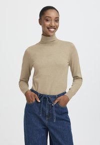 Donna che sorride in un maglione beige a collo alto e pantaloni di jeans blu a vita alta con cintura annodata, mani in tasca contro uno sfondo bianco.