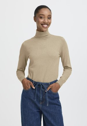 Donna che sorride in un maglione beige a collo alto e pantaloni di jeans blu a vita alta con cintura annodata, mani in tasca contro uno sfondo bianco.