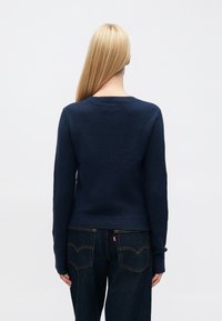 Tengerészkék kötött pulóver hosszú ujjakkal, kerek nyakkivágással és bordázott mandzsettákkal. Sötét farmernadrággal párosítva, amely látható varrással és logóval rendelkezik.