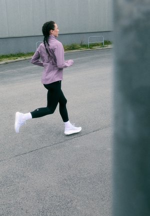 Violet bėgimo striukė, juodos leginsai ir baltos bėgimo bateliai. Pleurinta šukuosena, judesyje pozicija lauko bėgimo take su pilku fono.