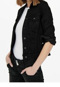Mustan denim-takki, jossa on etupainike, läpälliset taskut ja käännetyt hihat. Päällä valkoinen paita ja mustat farkut.
