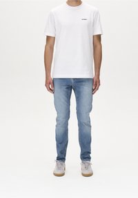 Bílé bavlněné tričko s kulatým výstřihem a malým černým logem, v kombinaci se světle modrými džíny a šedými teniskami s béžovými podrážkami.