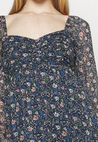 Blommig klänning med en marinblå bas och mönster av rosa och blå blommor, rynkad byst, fyrkantig halsringning och långa genomskinliga ärmar.